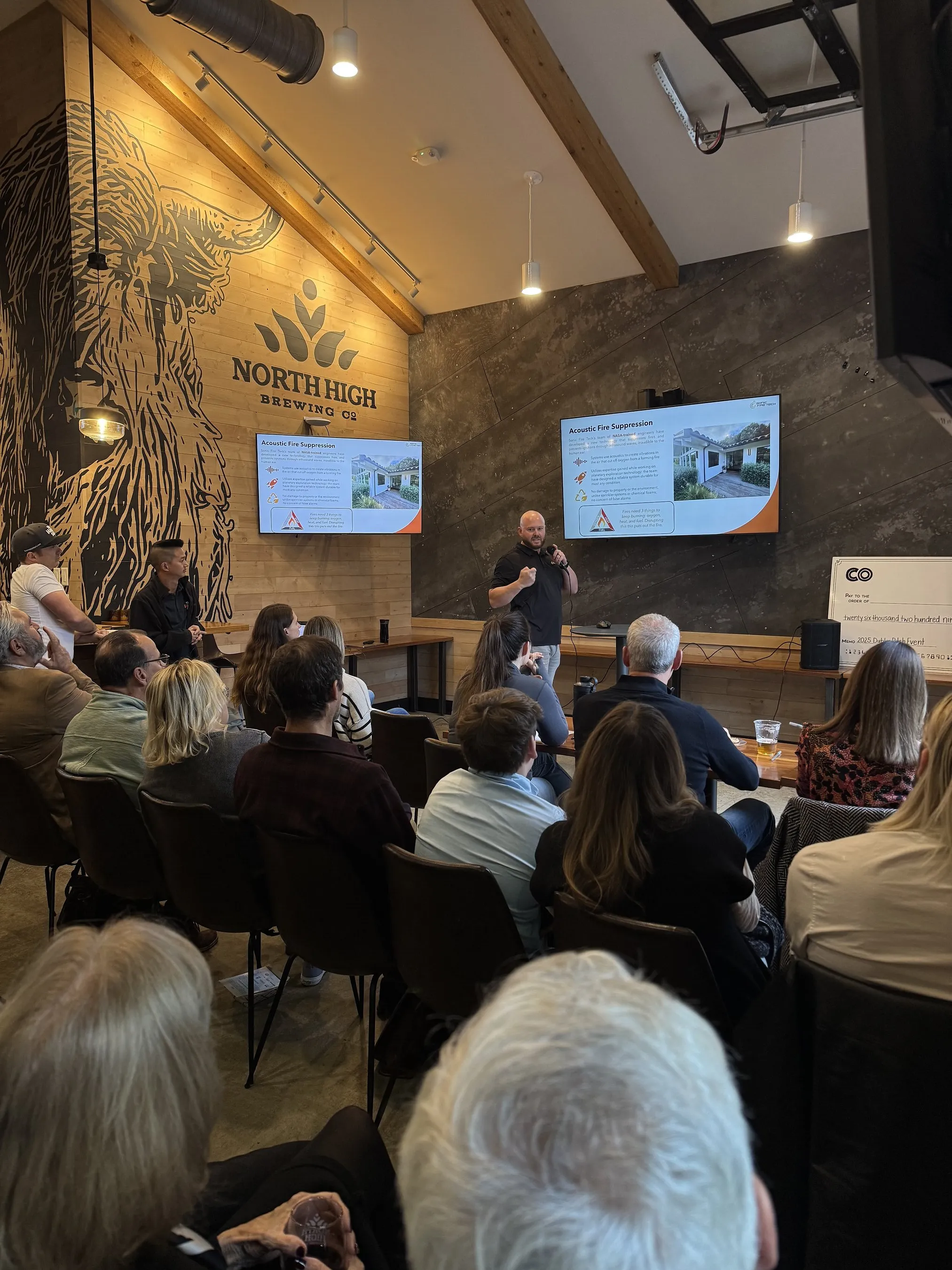Audience members watch a presenter speaking beside slides in a brewery meeting room.