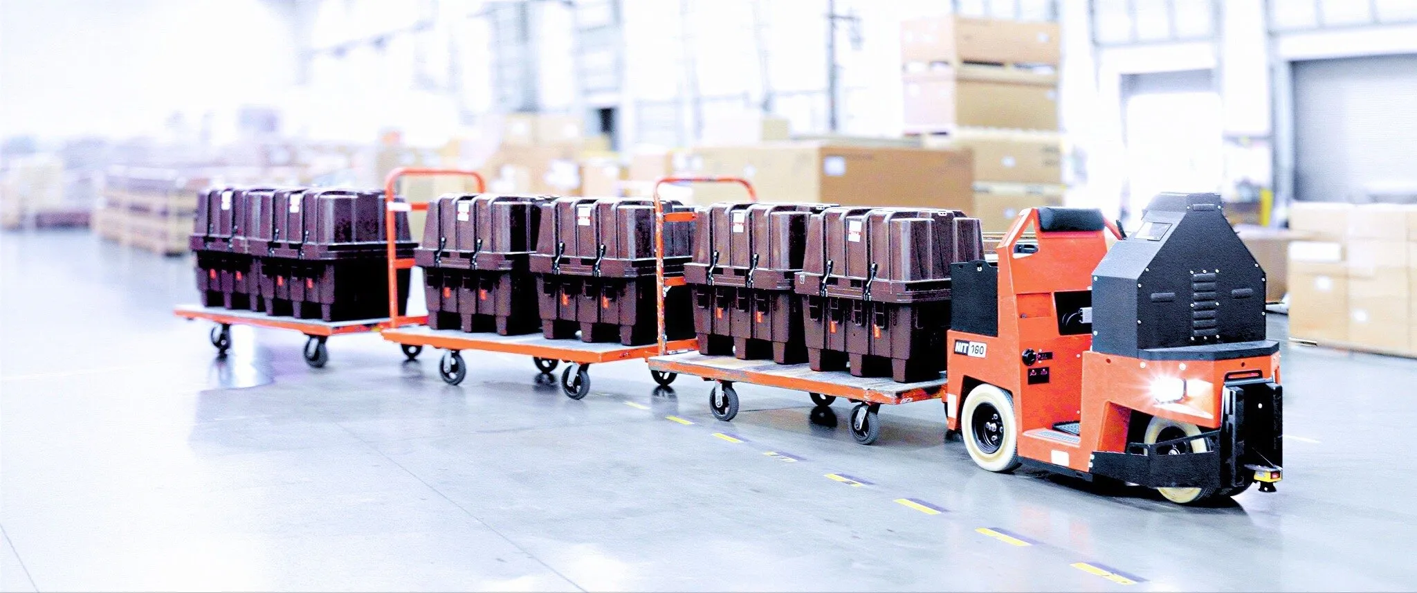 An orange autonomous tugger pulls carts loaded with black bins through a warehouse.