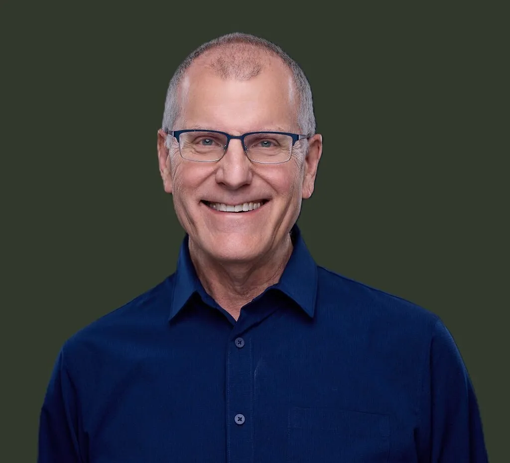 Greg DuPey smiling in glasses and a blue collared shirt against a dark background.