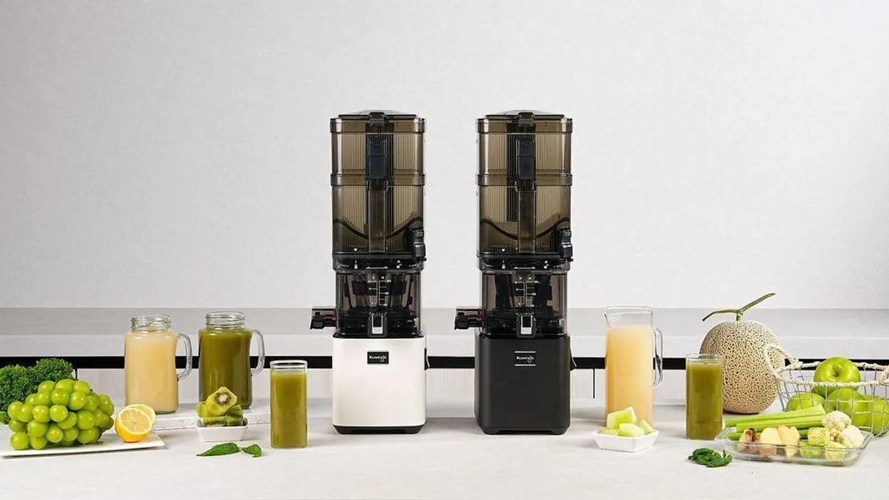 Two Kuvings juicers with glasses of green and orange juice, surrounded by fresh fruit and vegetables.