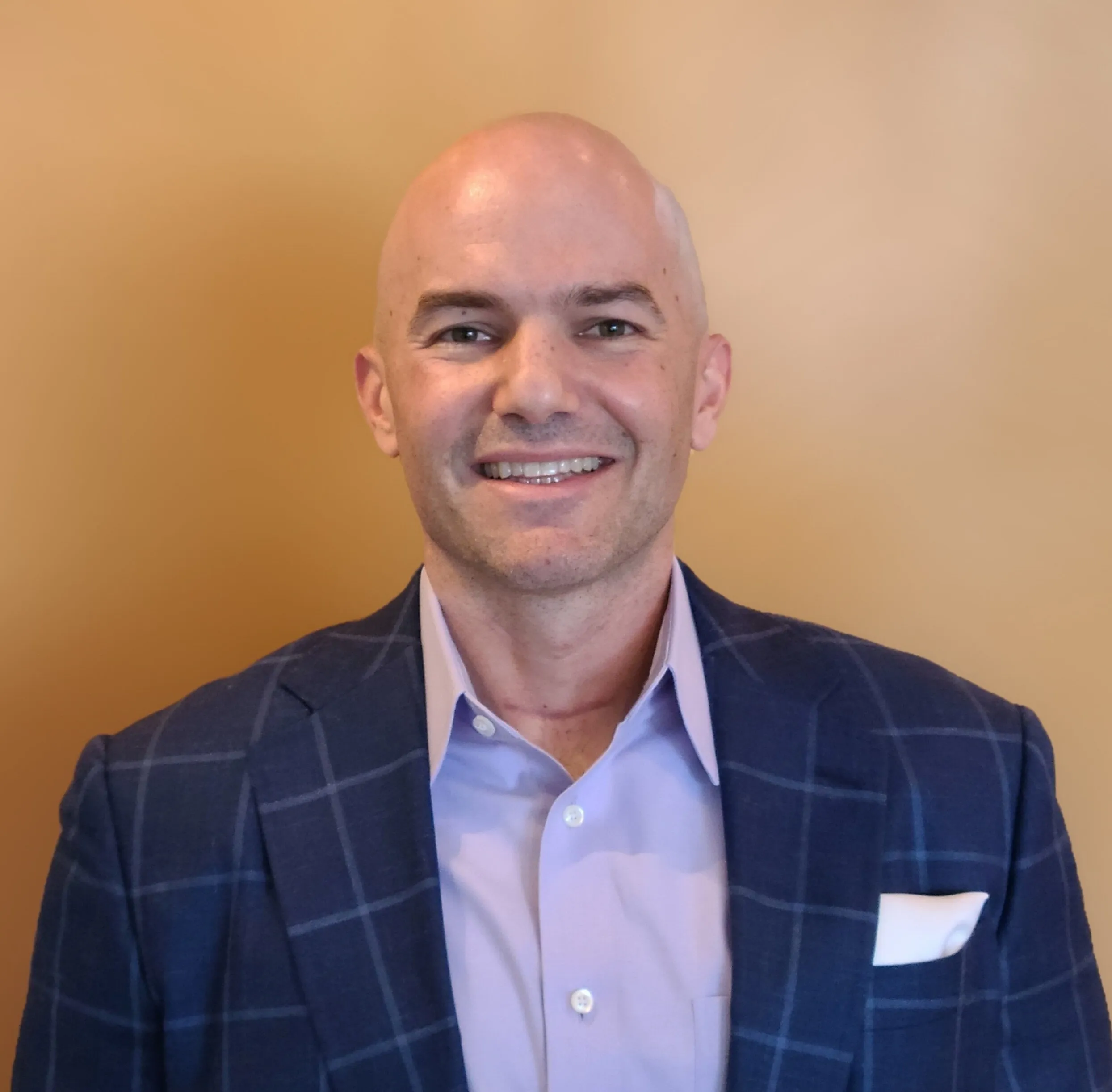 Mike Solow smiling in a navy check blazer and lavender shirt against a tan background.
