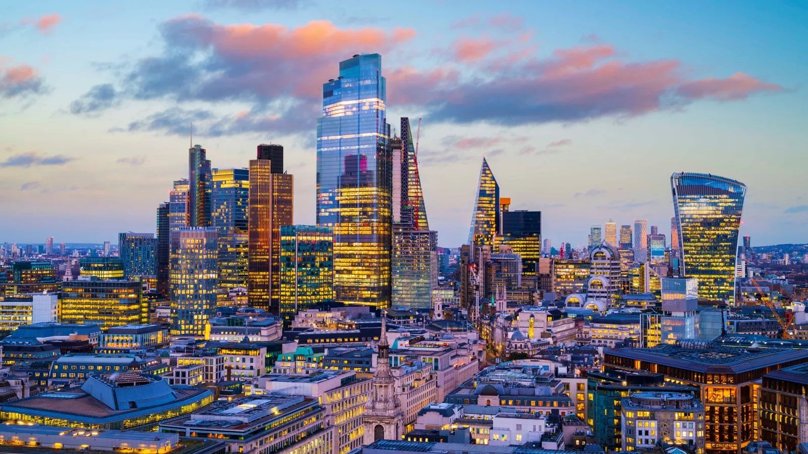 Office towers of the City of London lit up at dusk, seen from above the rooftops