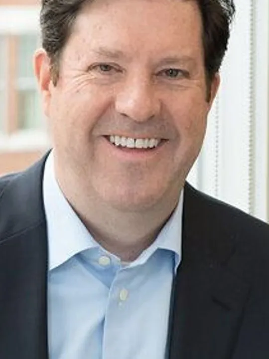 A smiling person in a dark suit and light blue shirt stands by a window.